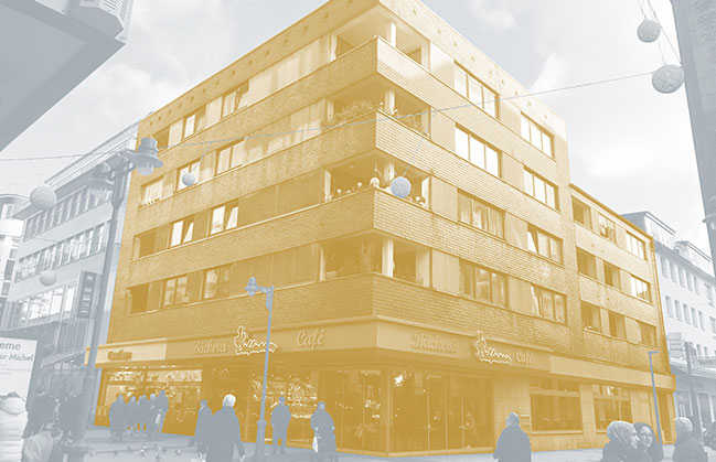 Das Bild zeigt ein gelb hervorgehobes Wohn- und Geschäftshaus im Zentrum Wuppertals. Es ist umgeben von einer aktiven Fußgängerstraße und grenzt an zwei weiteren Gebäuden.